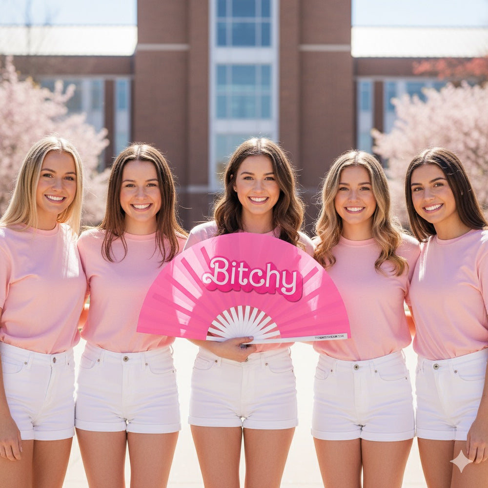 Group of women holding a pink hand fan with 'Bitchy' text in a casual setting