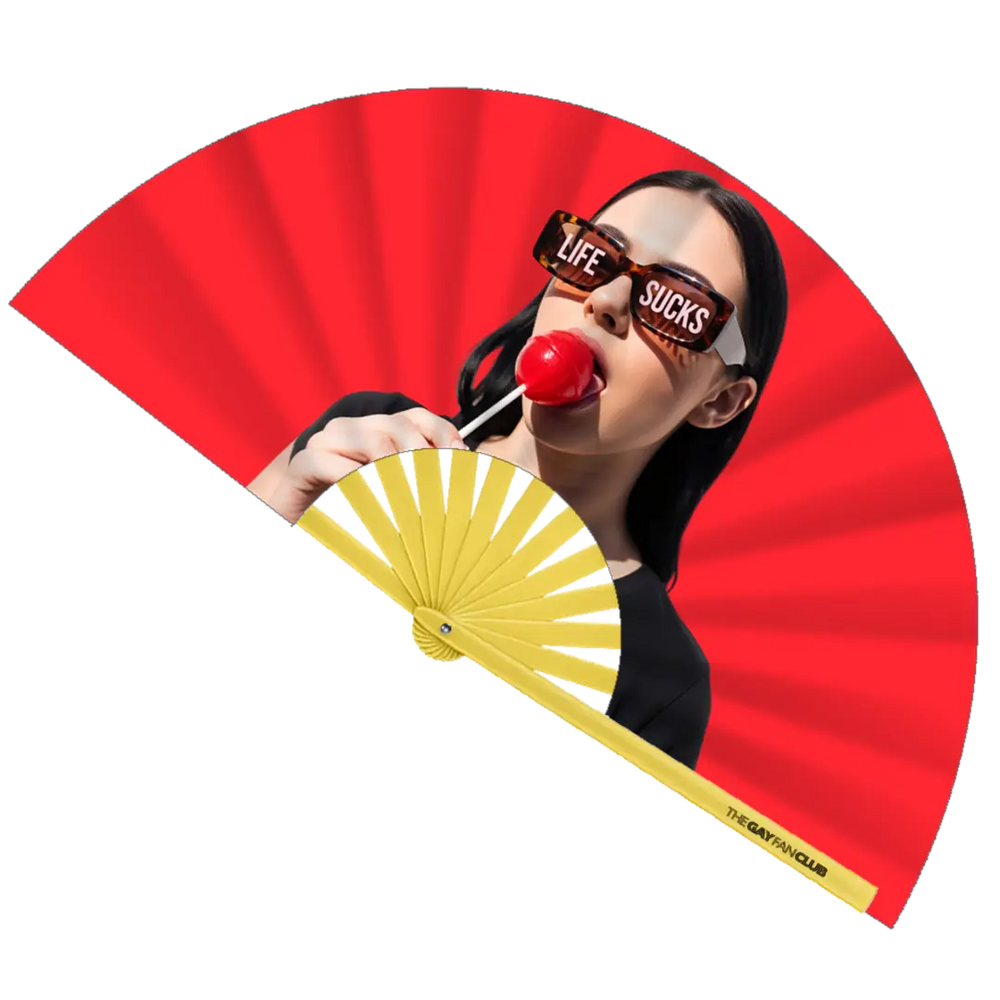 Best Life Fan fully open showing a woman in tortoise-shell sunglasses with "LIFE SUCKS" text on the lenses, holding a red lollipop, set against a vibrant red background and yellow bamboo ribs.