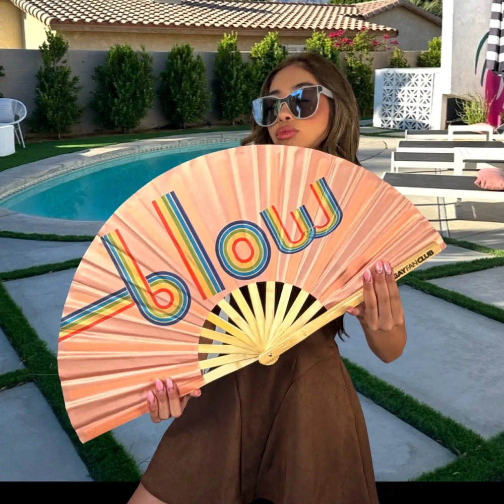 Person holding a colorful 'Blow' fan by a pool with a scenic background