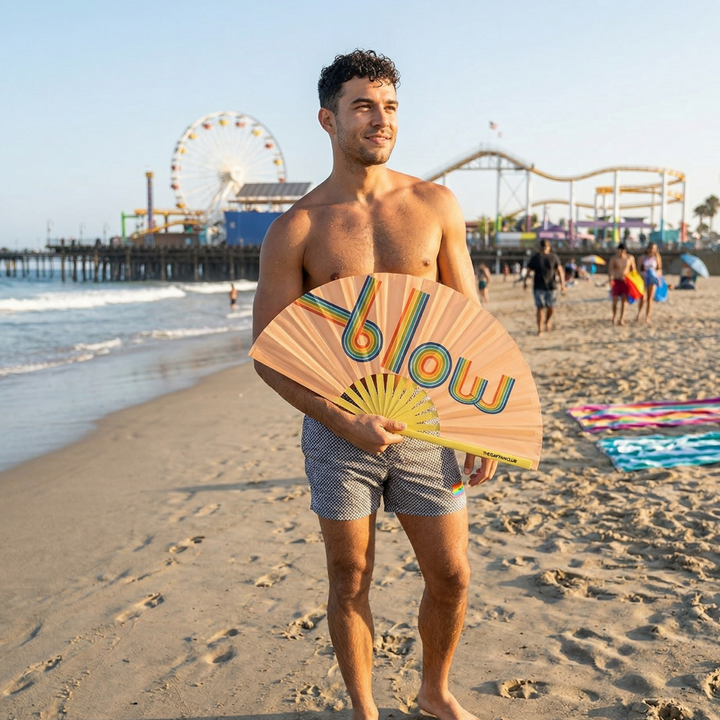 Man holding a colorful "blow" bamboo clack fan on a beach with a Ferris wheel and pier in the background