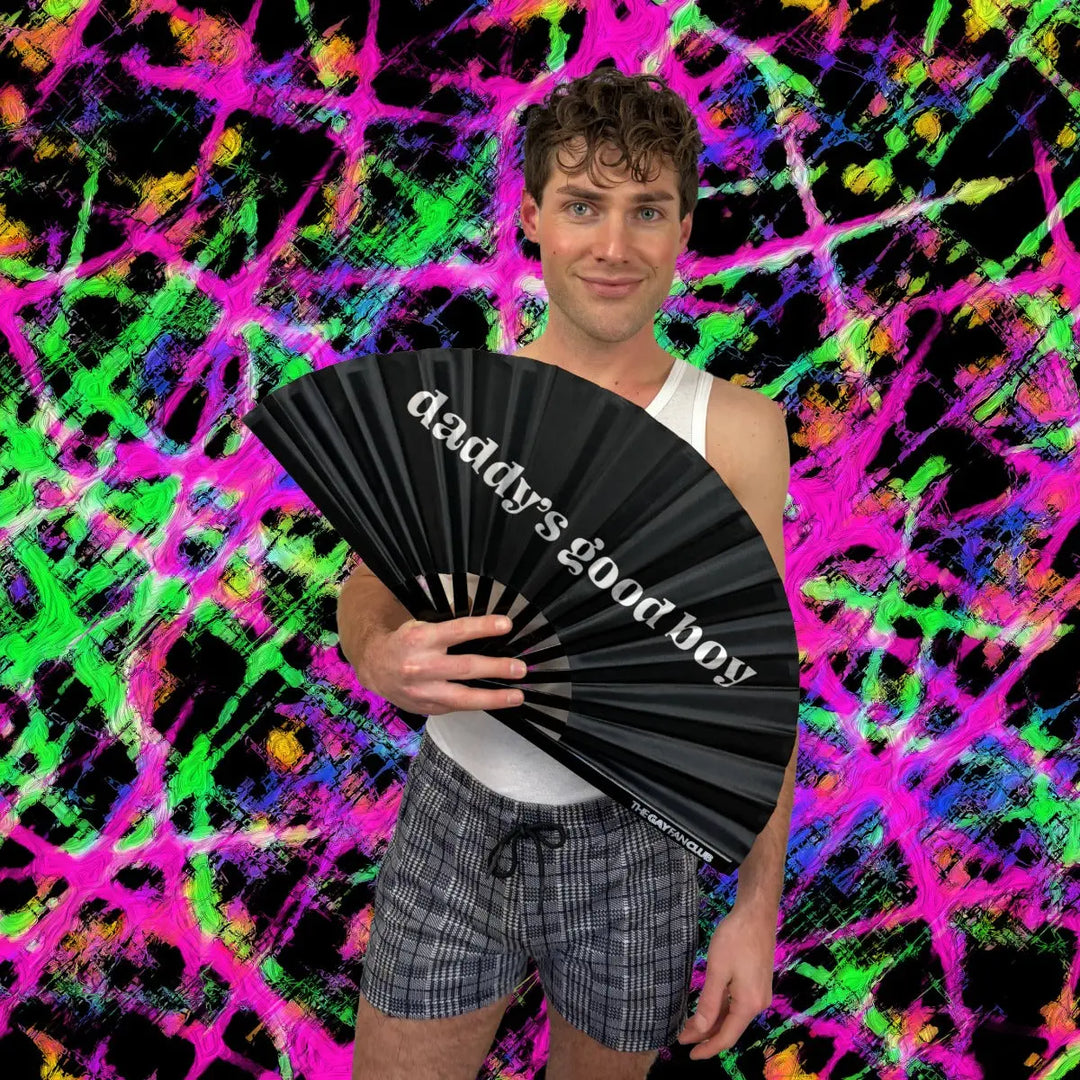 Man holding a black fan with 'daddy's good boy' text against a colorful abstract background