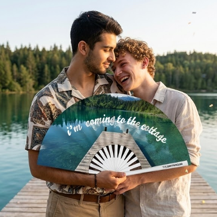 A lgbt couple holding the "Coming to the Cottage" fan at a book convention, highlighting the oversized 13-inch frame.