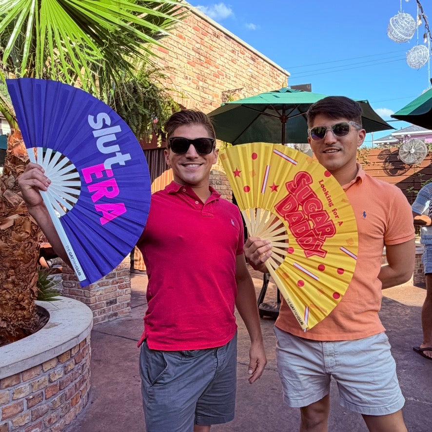 Gay twins holding two funny statement clack fans from The Gay Fan Club the "looking for a sugar daddy" and "Slut Era" fans