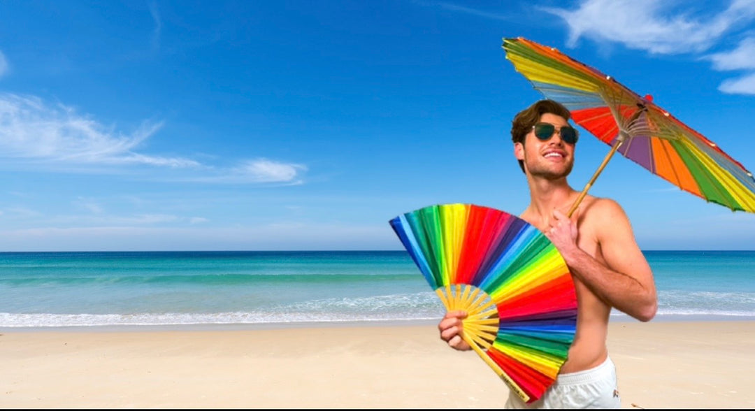 Man on a beach holding a colorful umbrella and rainbow bamboo clack fan from The Gay Fan Club with blue sky and ocean in the background