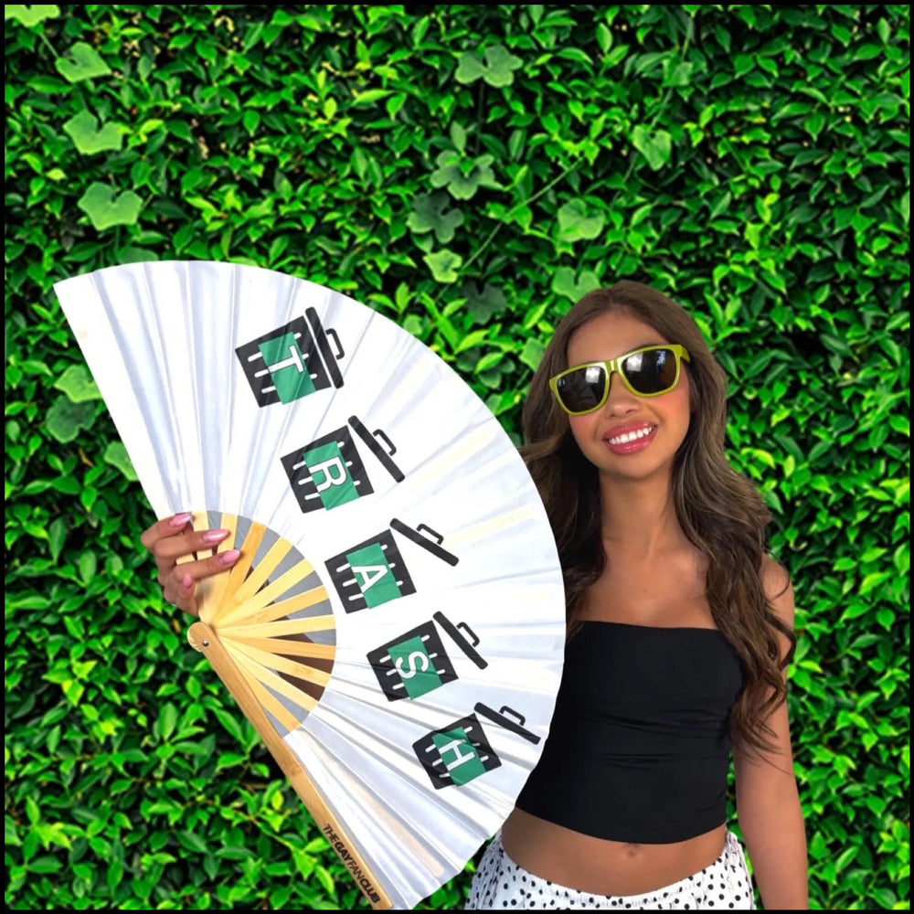 Woman holding a white fan with 'TRASH' branding against a green hedge background